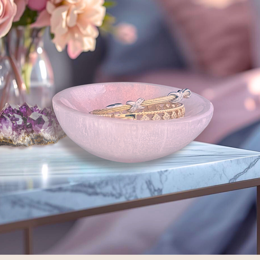 Red blush selenite crystal bowl with gold bracelets on a marble table with flowers and crystals in the backgrouncd