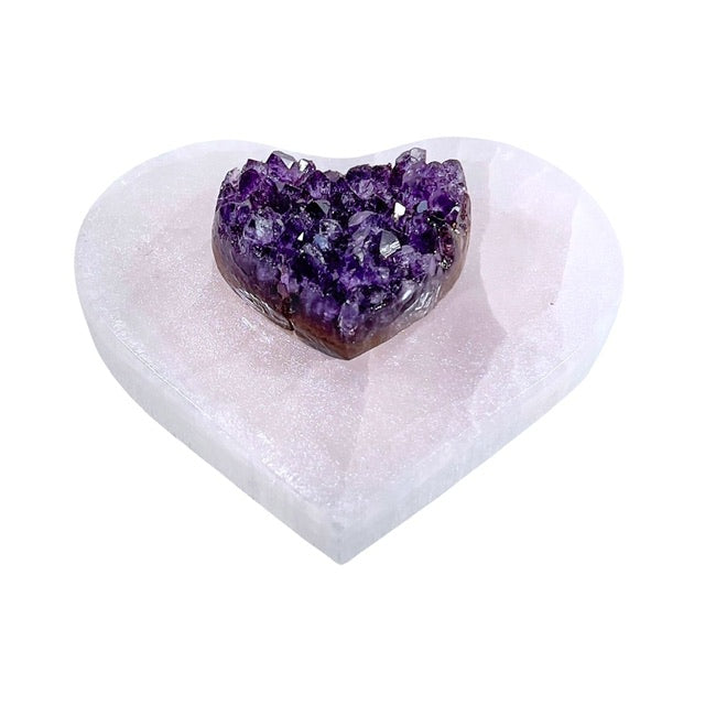 A tilted view of a stunning selenite heart charging plate featuring a mesmerizing pink shimmery finish. Adorned with a beautiful AAA-grade, 15mm amethyst heart at the center,  the perfect crystal for cleansing and charging your crystals.