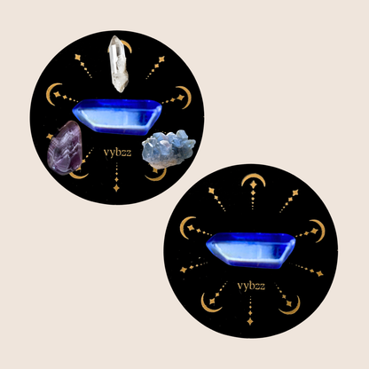Top view of two black round aromatherapy candle lids with blue crystals and celestial gold patterns, and assorted healing stones.